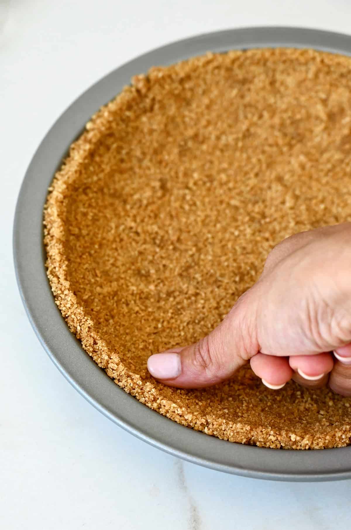 A hand presses a crushed graham cracker, melted butter and brown sugar mixture into a pie plate to make a graham cracker crust.
