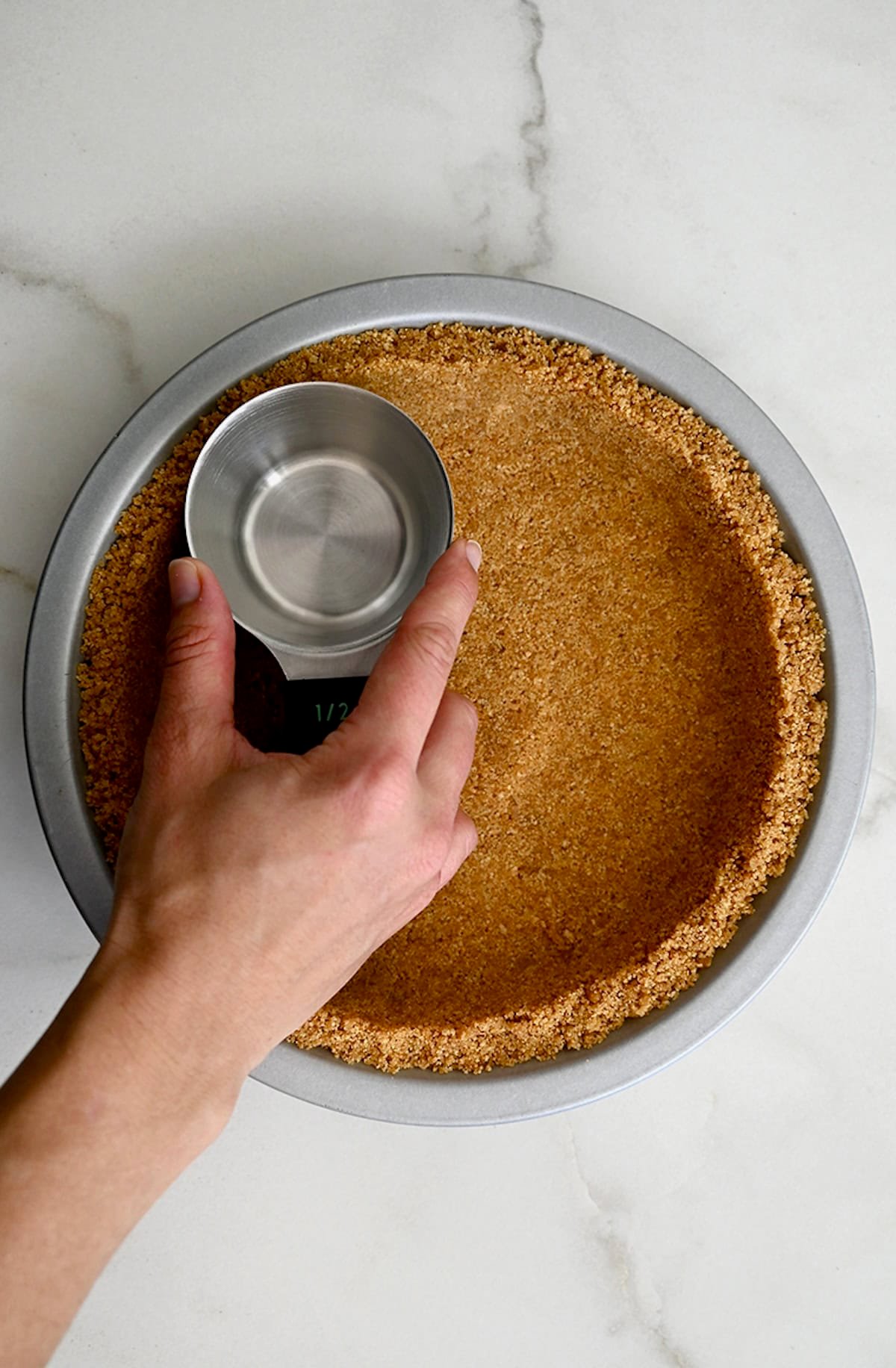 A hand holding a measuring cup presses a graham cracker crumb mixture into a pie pan to form a graham cracker crust.
