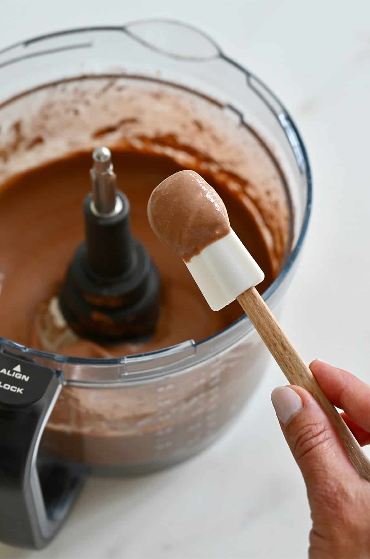 A white spatula with mousse on it with a food processor bowl in the background.