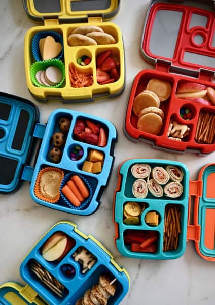 Four Bentgo lunchboxes with fresh fruits and veggies, pinwheel sandwiches, mini pancakes, protein balls and French toast skewers.
