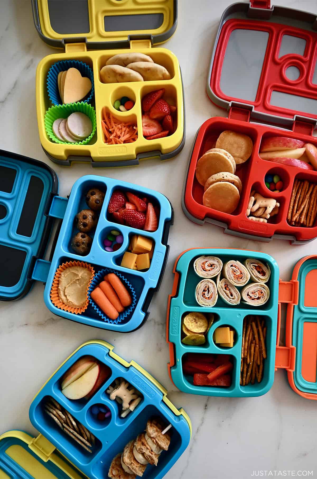 Four Bentgo lunchboxes with fresh fruits and veggies, pinwheel sandwiches, mini pancakes, protein balls and French toast skewers.