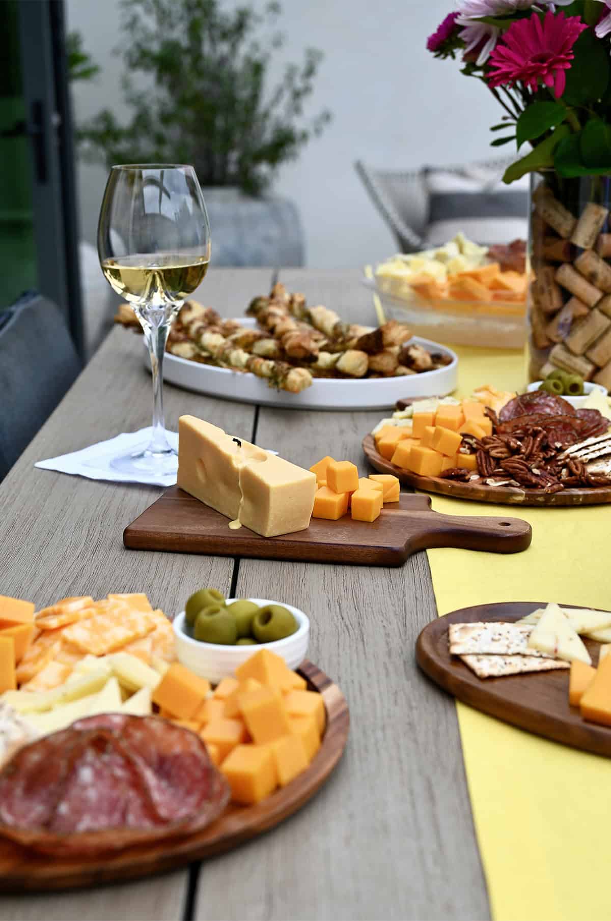 Wine and cheese party spread on a wooden table, featuring assorted cheeses, crackers, cured meats, fresh fruit, jam, and wine glasses, with decorative touches like small cheese boards and cheese candle centerpiece. 
