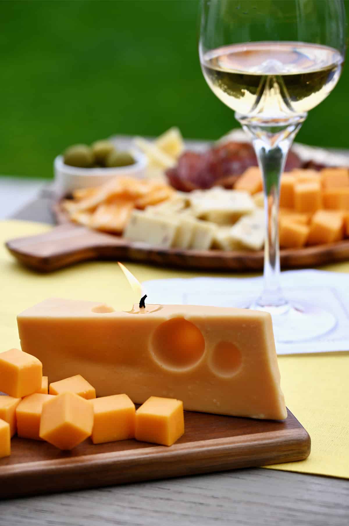 Cheese candle on a charcuterie board with cubed cheddar cheese. A glass of white wine is nearby.