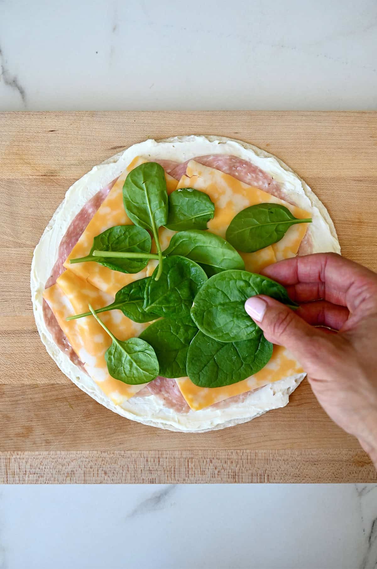 Thinly sliced salami, Colby Jack cheese and spinach are being layered atop a tortilla with cream cheese spread.