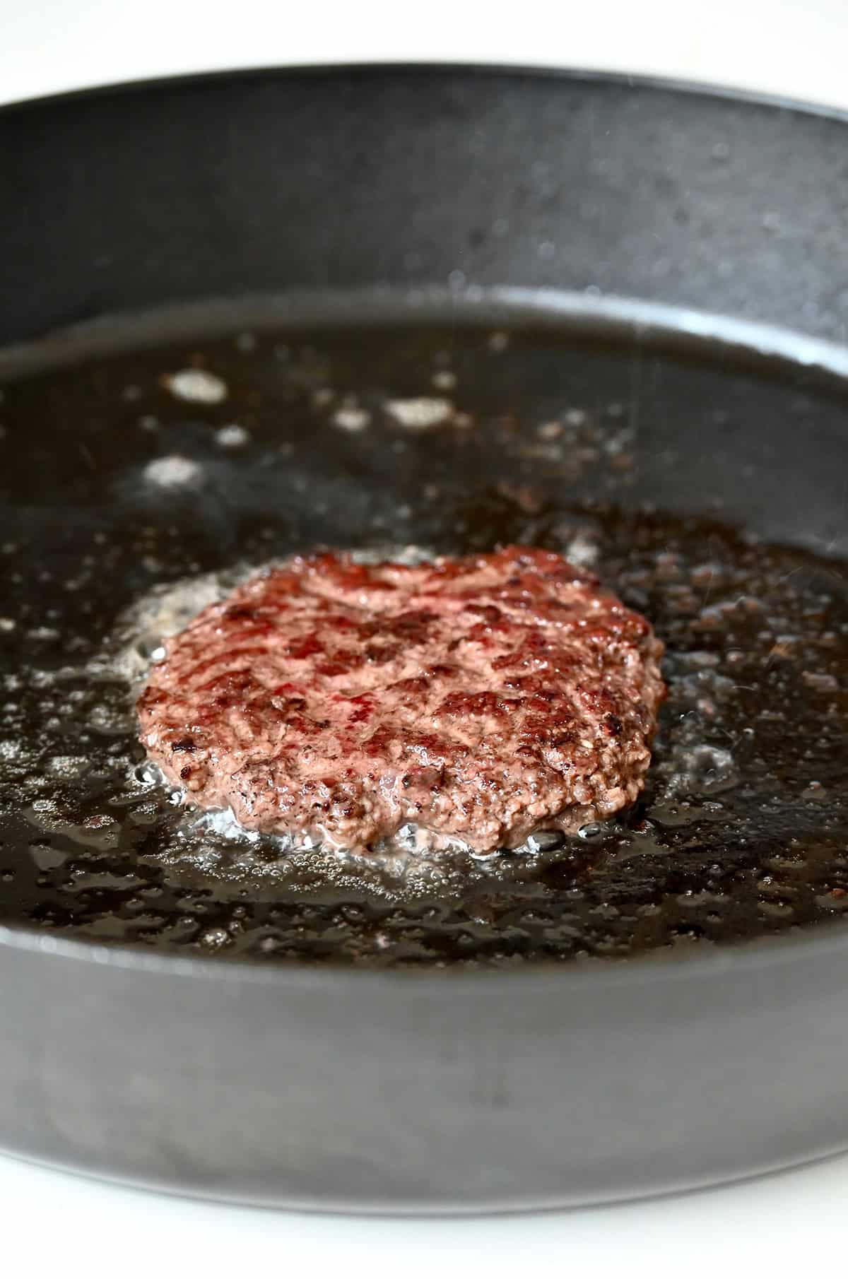 A slightly crispy, browned burger patty in a hot skillet with oil.