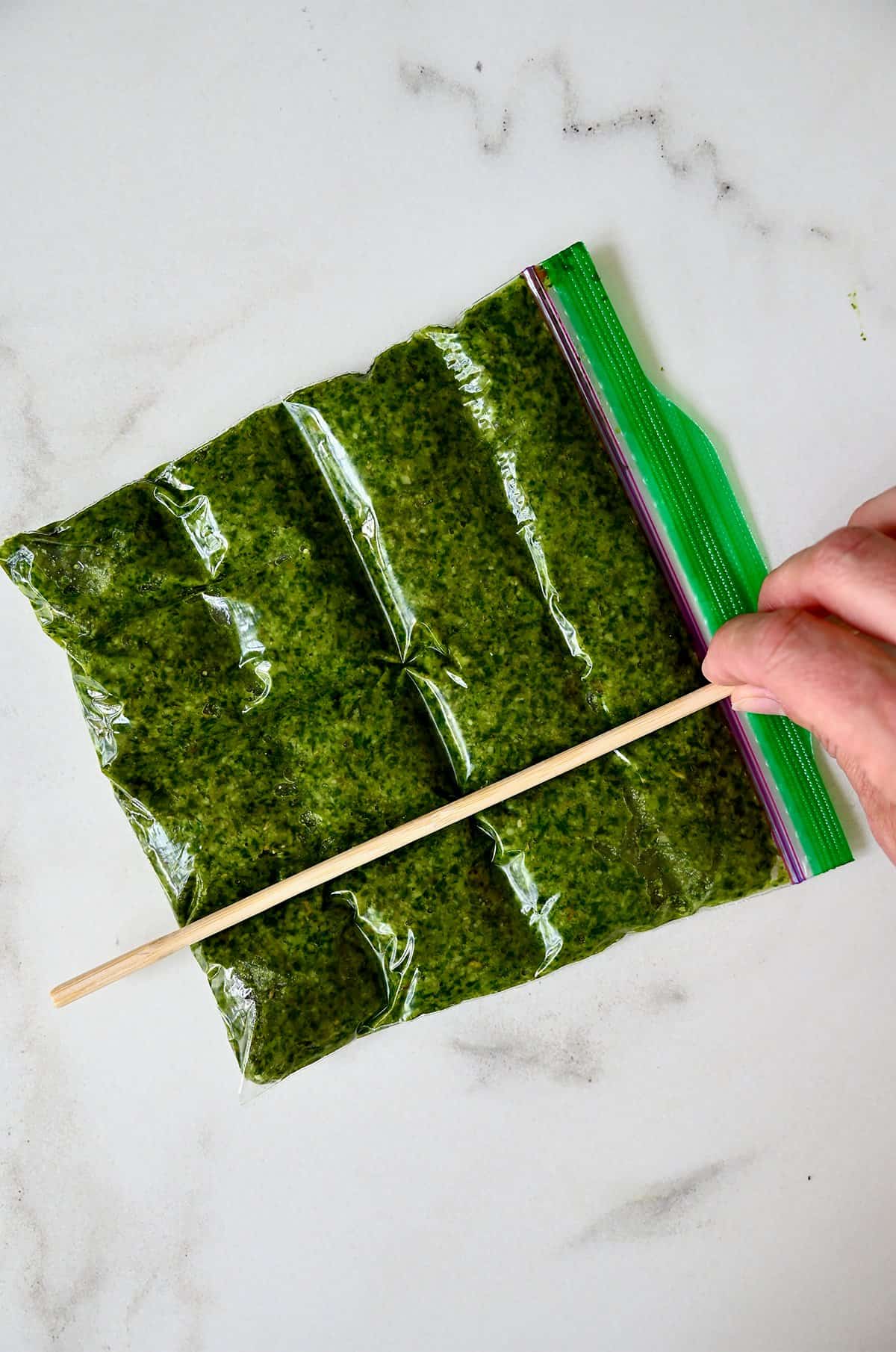 A freezer-safe plastic bag containing a layer of pesto that has been scored with a wooden stick to create individual portions for freezing.