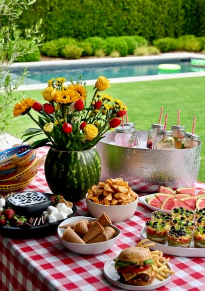 A colorful summer picnic party table set with barbecue food, a watermelon flower centerpiece, and a metal drink bucket filled with ice and cold beverages.