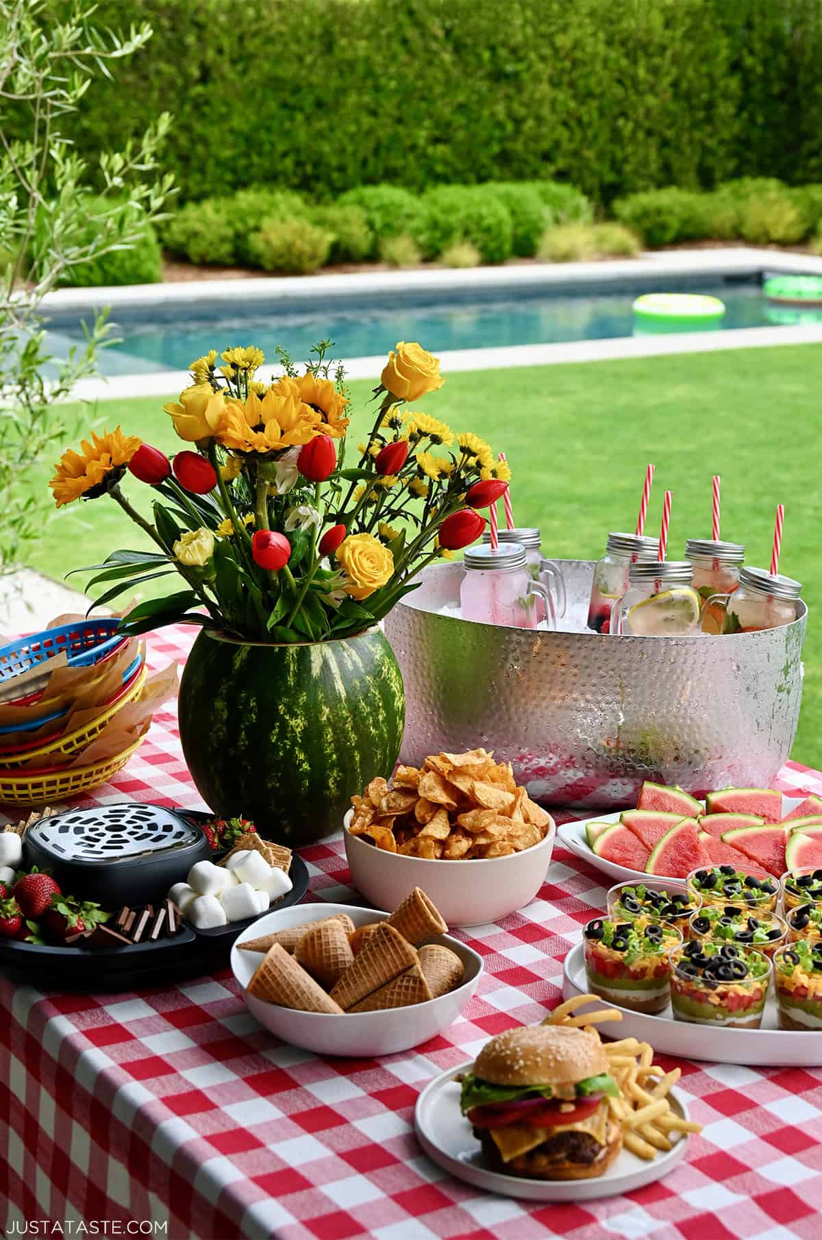 A colorful summer picnic party table set with barbecue food, a watermelon flower centerpiece, and a metal drink bucket filled with ice and cold beverages.