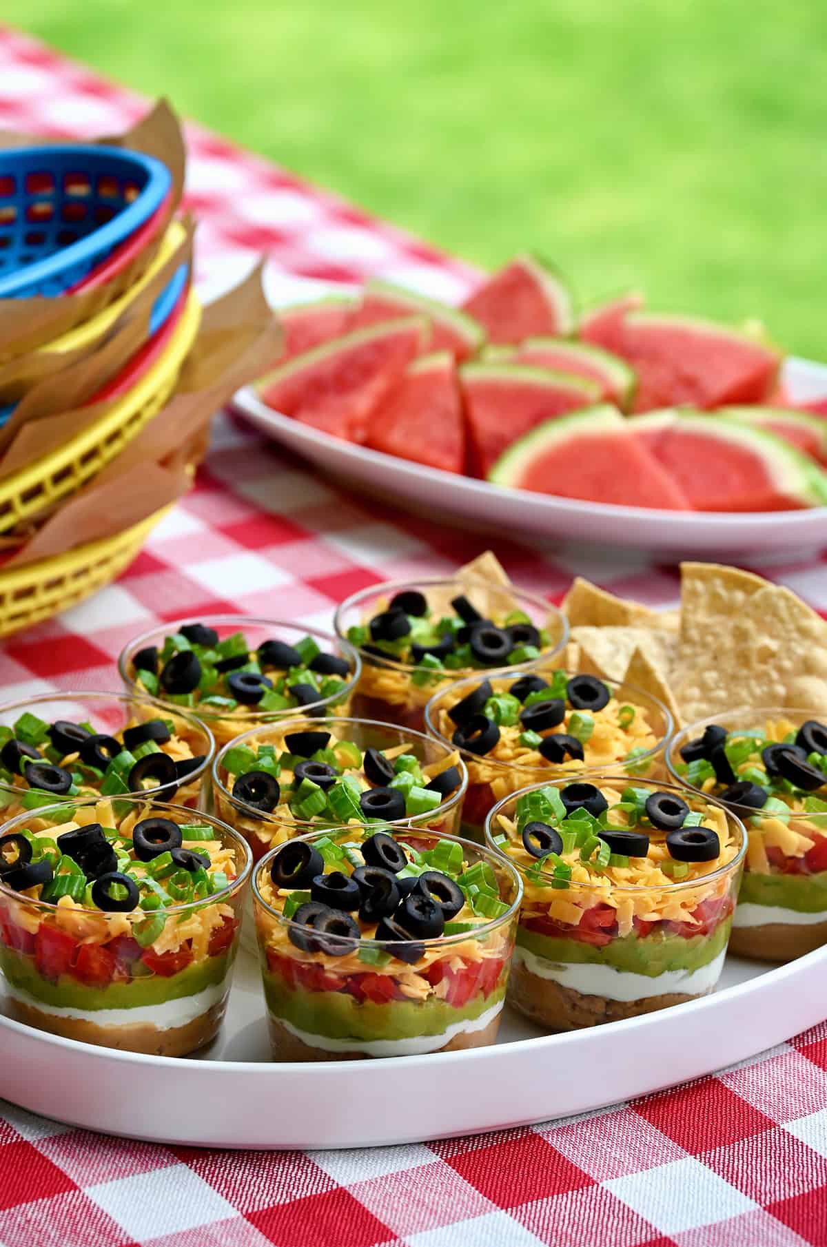 Individual 7-layer dips served in clear mini cups on a red and white checkered tablecloth, perfect for summer parties or BBQs.