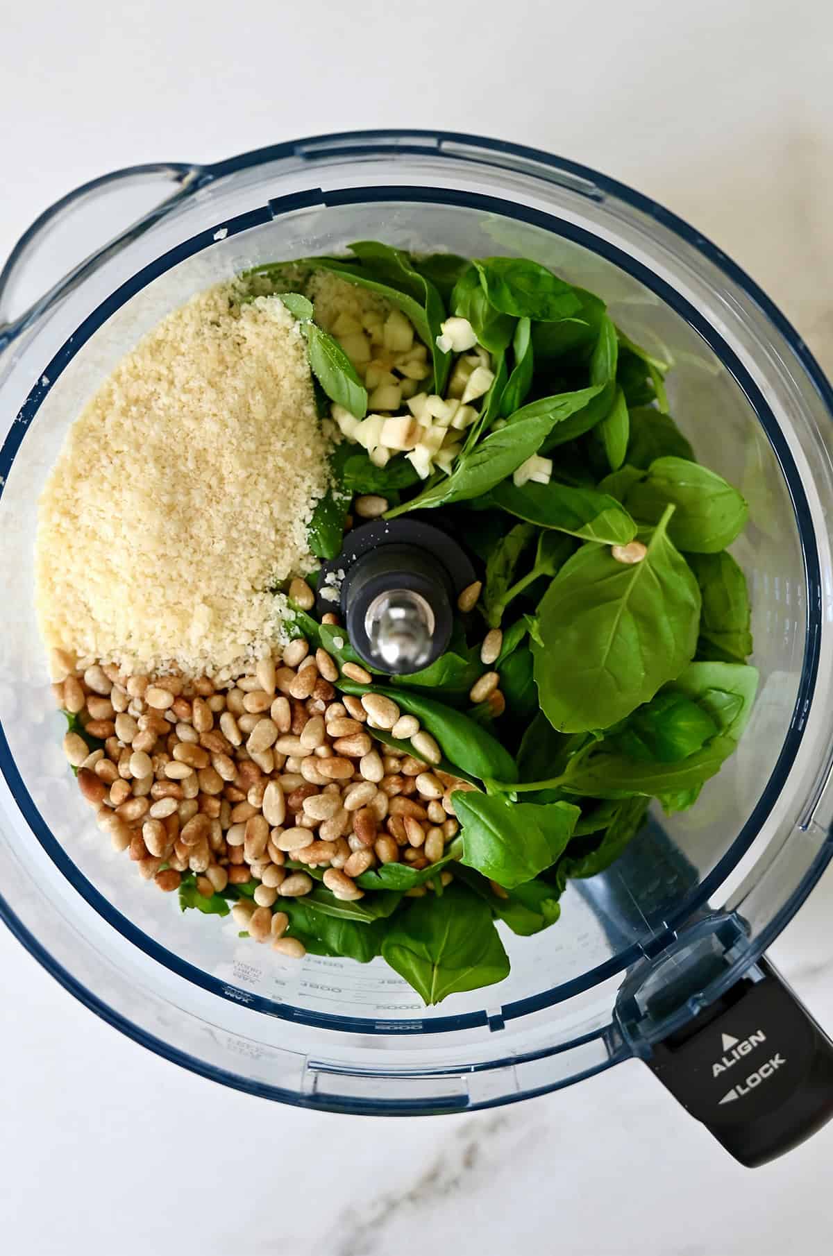 A food processor containing pine nuts, fresh basil leaves, garlic, salt, pepper and grated Parmesan cheese.