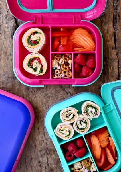 Two lunchboxes containing pinwheel sandwiches, fresh fruit and veggies, and trail mix.