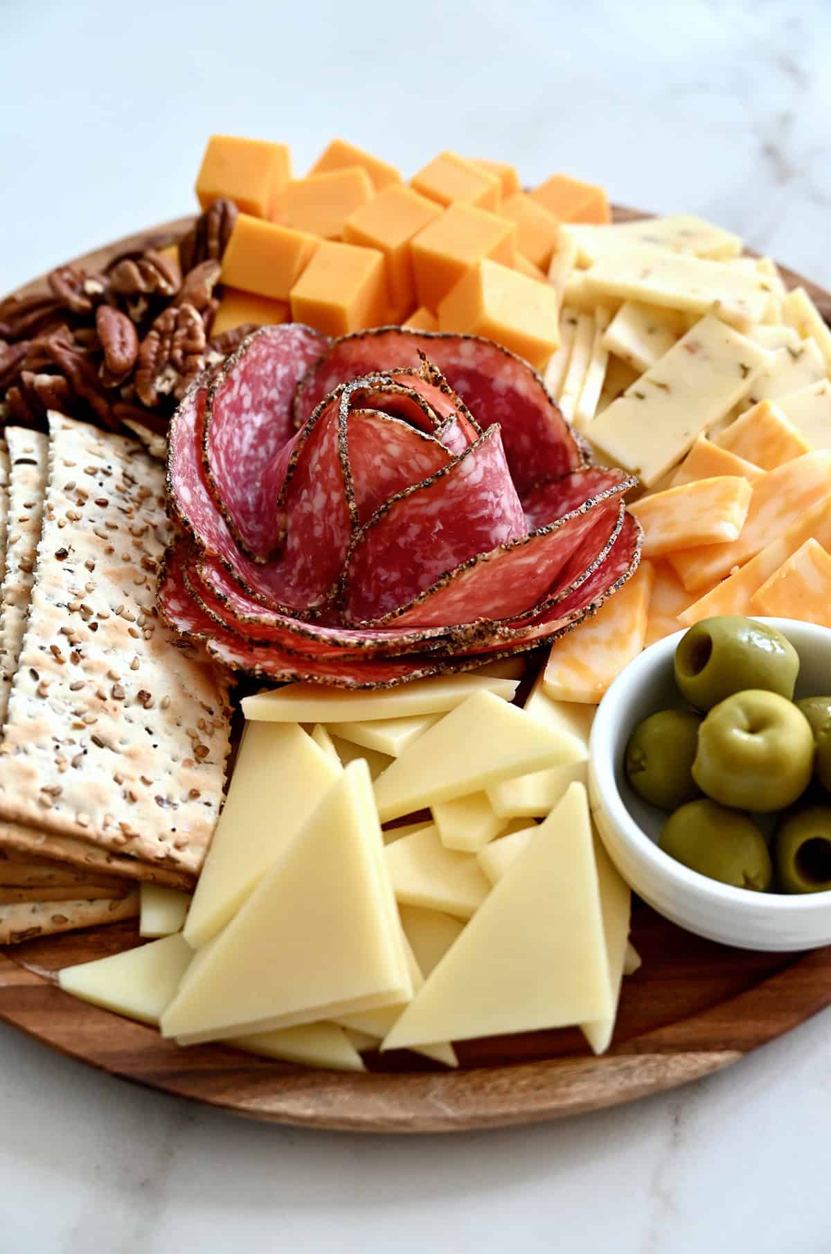 Salami rose on a charcuterie board surrounded by a variety of cheeses, crackers and olives. 