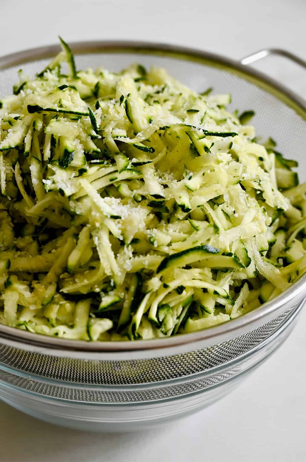 Shredded zucchini with salt in a sieve over a bowl.