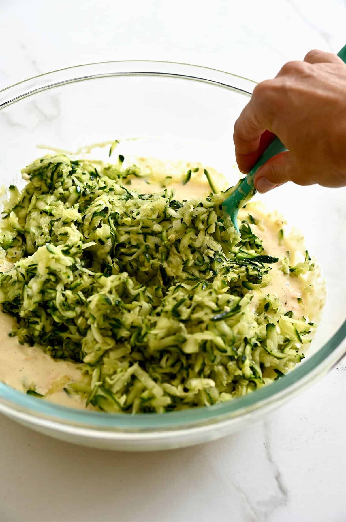 Grated zucchini being stirred into the cheesy mixture with a spatula.