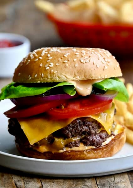 A homemade smash burger with double beef patties, a slice of cheddar cheese, tomatoes, lettuce and homemade burger sauce on a plate with French fries.