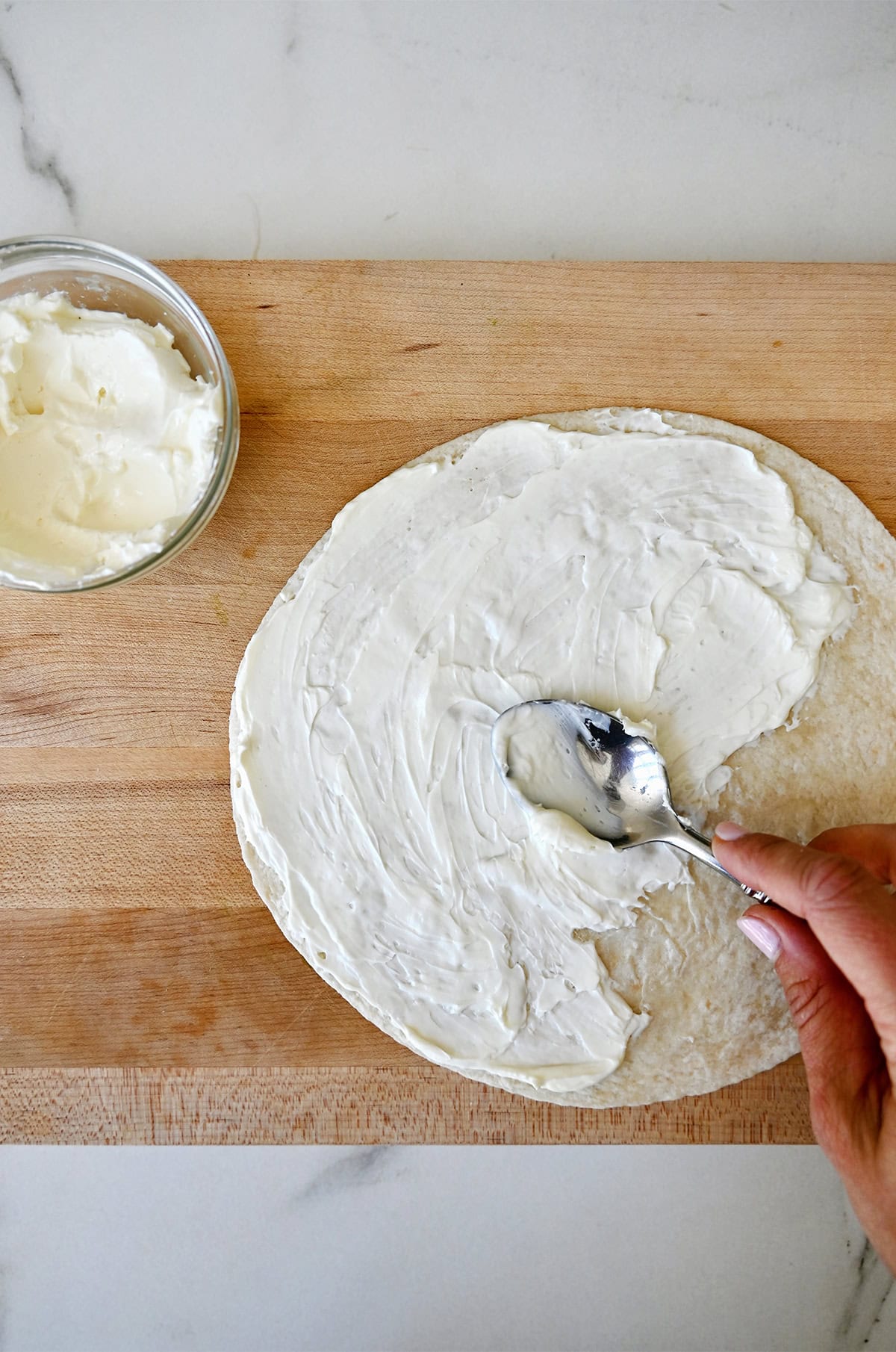 Spreading cream cheese all the way to the edges of a flour tortilla.
