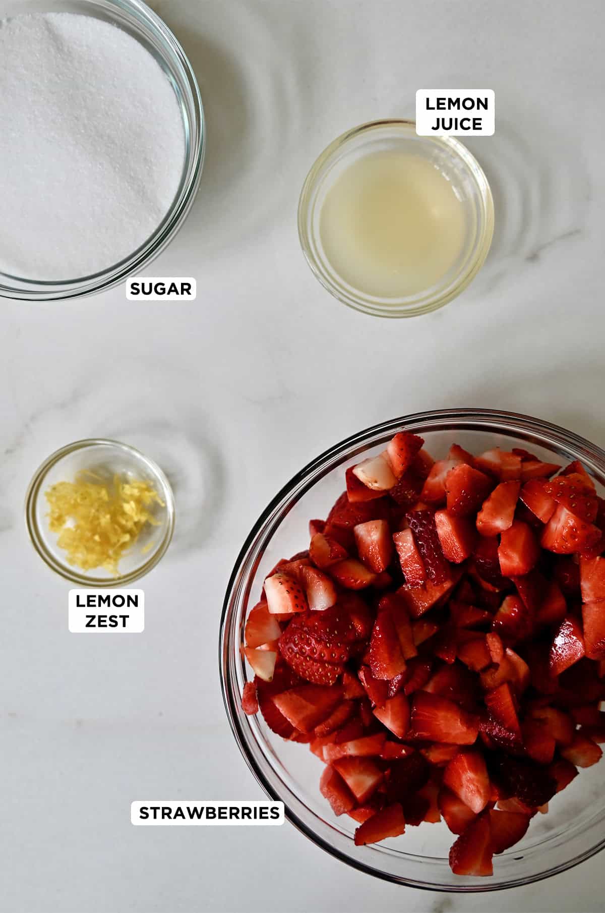 The four ingredients needed to make homemade strawberry sauce: fresh strawberries, white sugar, lemon juice and lemon zest.