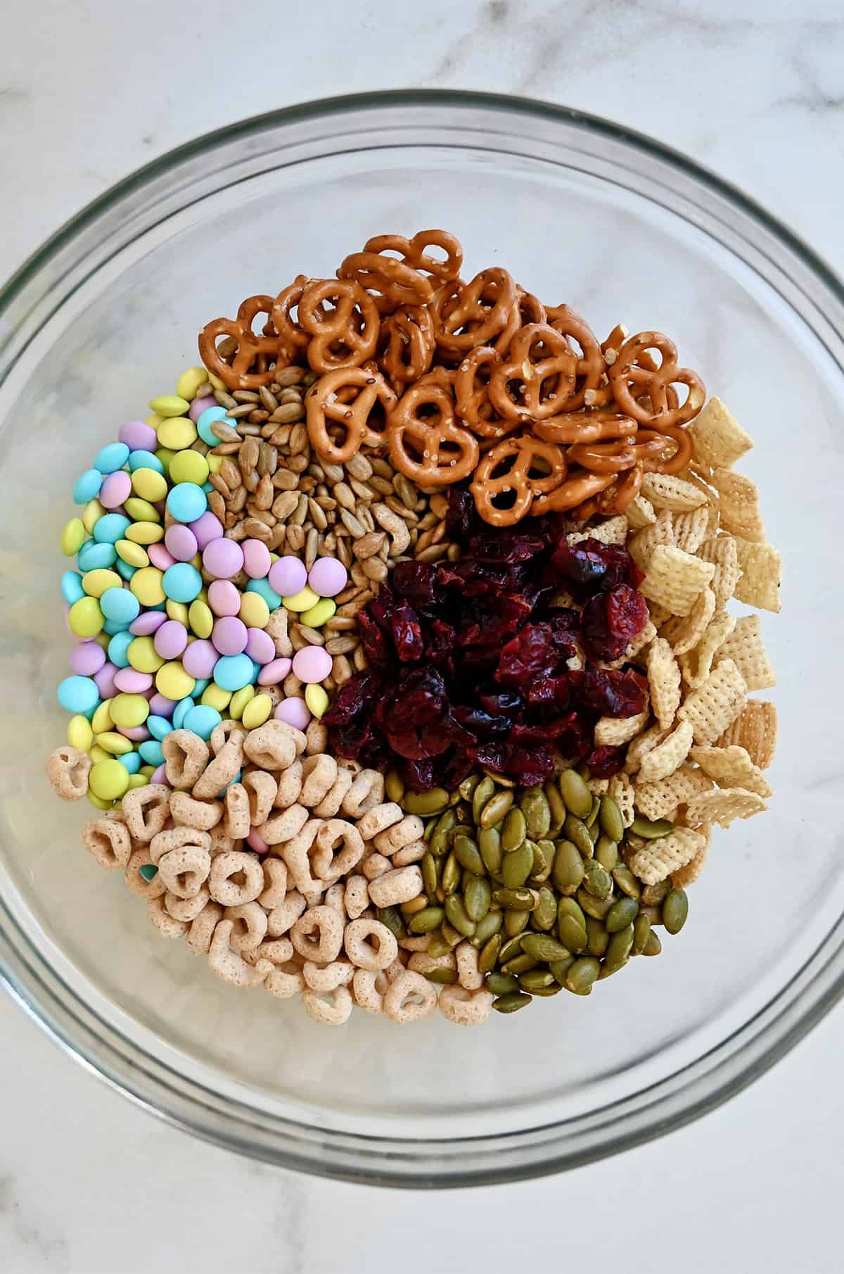 Pastel colored M&Ms, sunflower seeds, mini pretzels, Chex cereal, pumpkin seeds, Cheerios and dried cranberries in a bowl.