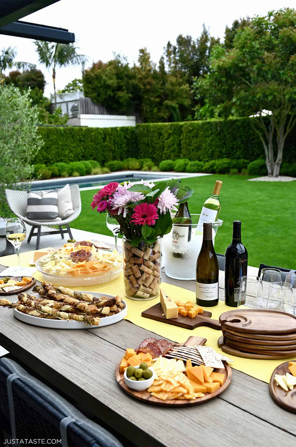 A table set for a wine and cheese party with red and white wines, a floral arrangement in a corkscrew vase, individual charcuterie boards and snacks.
