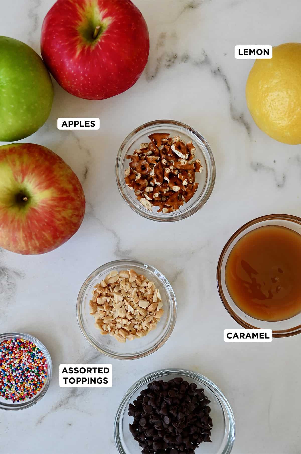 Ingredients to make apple nachos including apples, caramel sauce, sprinkles, crushed peanuts, crushed pretzels, mini chocolate chips, and a lemon to help prevent the apples from browning.