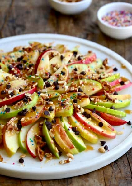 Caramel apple nachos starring thin slices of apples topped with a thick drizzle of caramel sauce, mini chocolate chips, chopped peanuts, chopped pretzels and rainbow sprinkles.