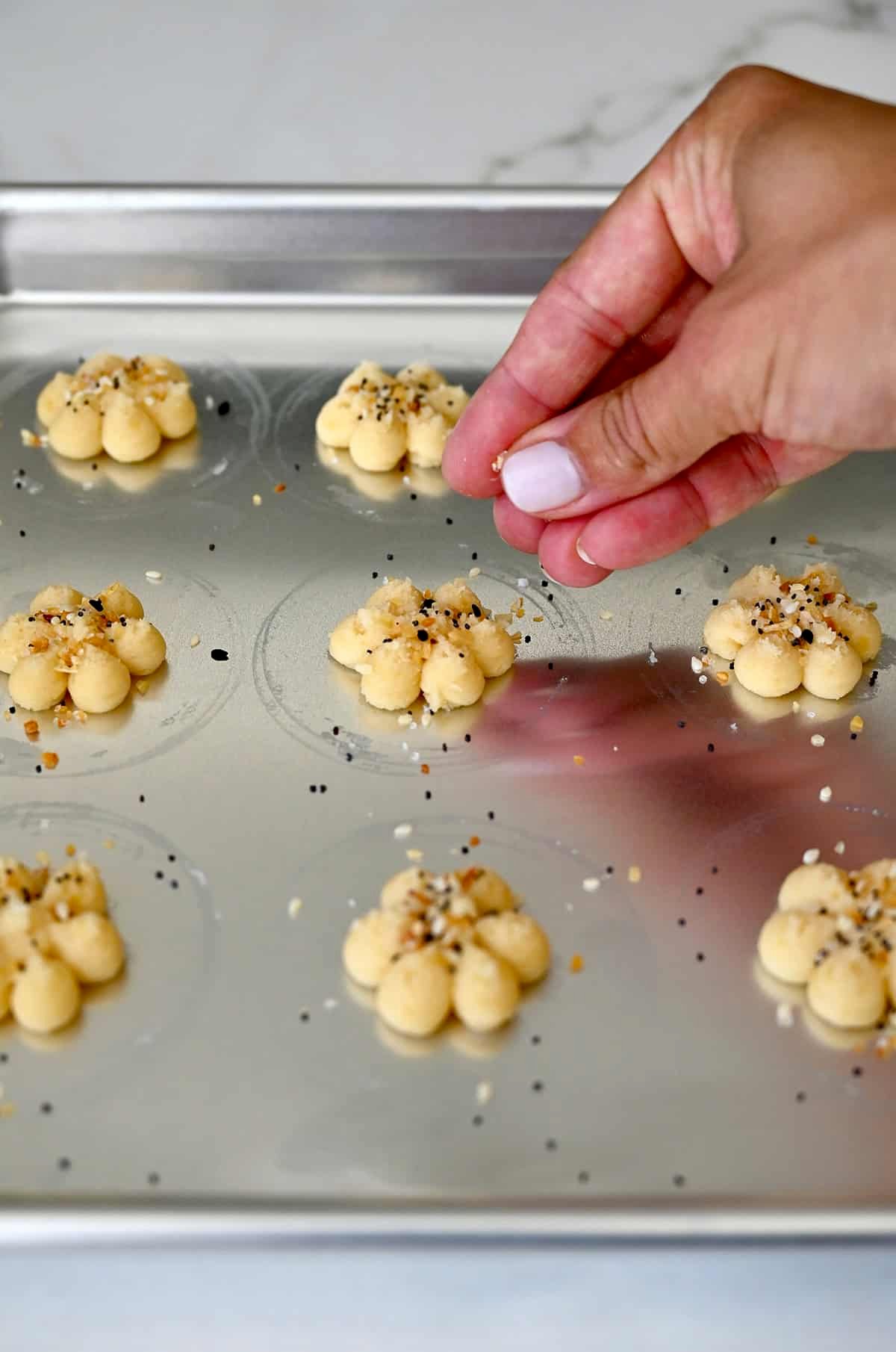 A hand sprinkles everything bagel seasoning atop cheddar spritz cookie dough.