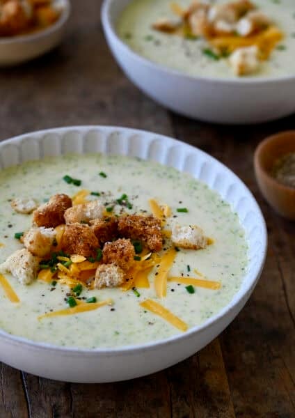A bowl containing creamy homemade broccoli cheddar soup topped with homemade croutons, chopped chives and shredded cheddar cheese.