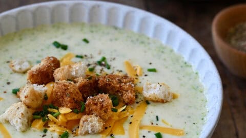 A bowl containing creamy homemade broccoli cheddar soup topped with homemade croutons, chopped chives and shredded cheddar cheese.