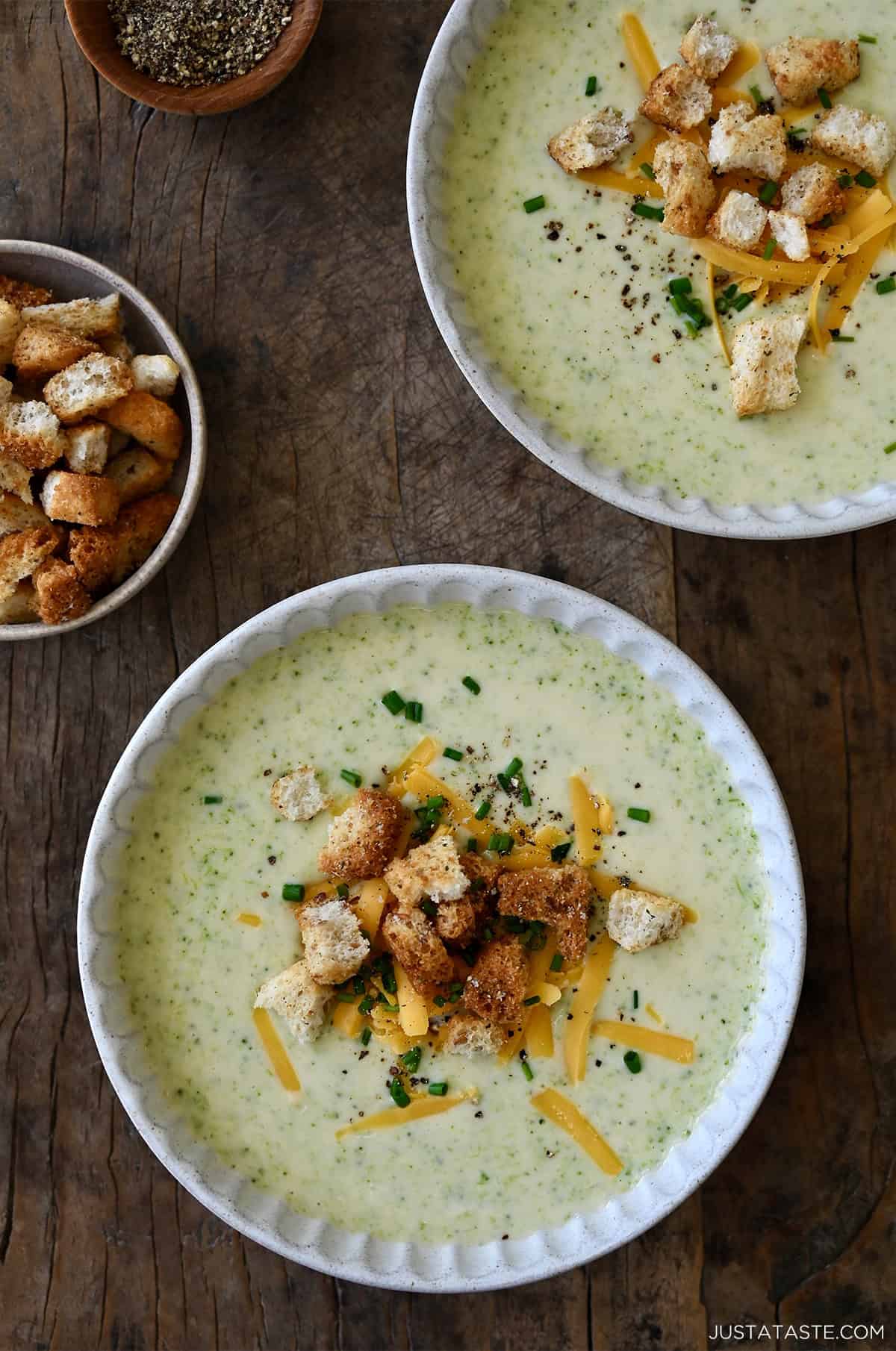 Two bowls containing creamy homemade broccoli cheddar soup topped with crunchy croutons, shredded cheddar cheese and chopped fresh chives.