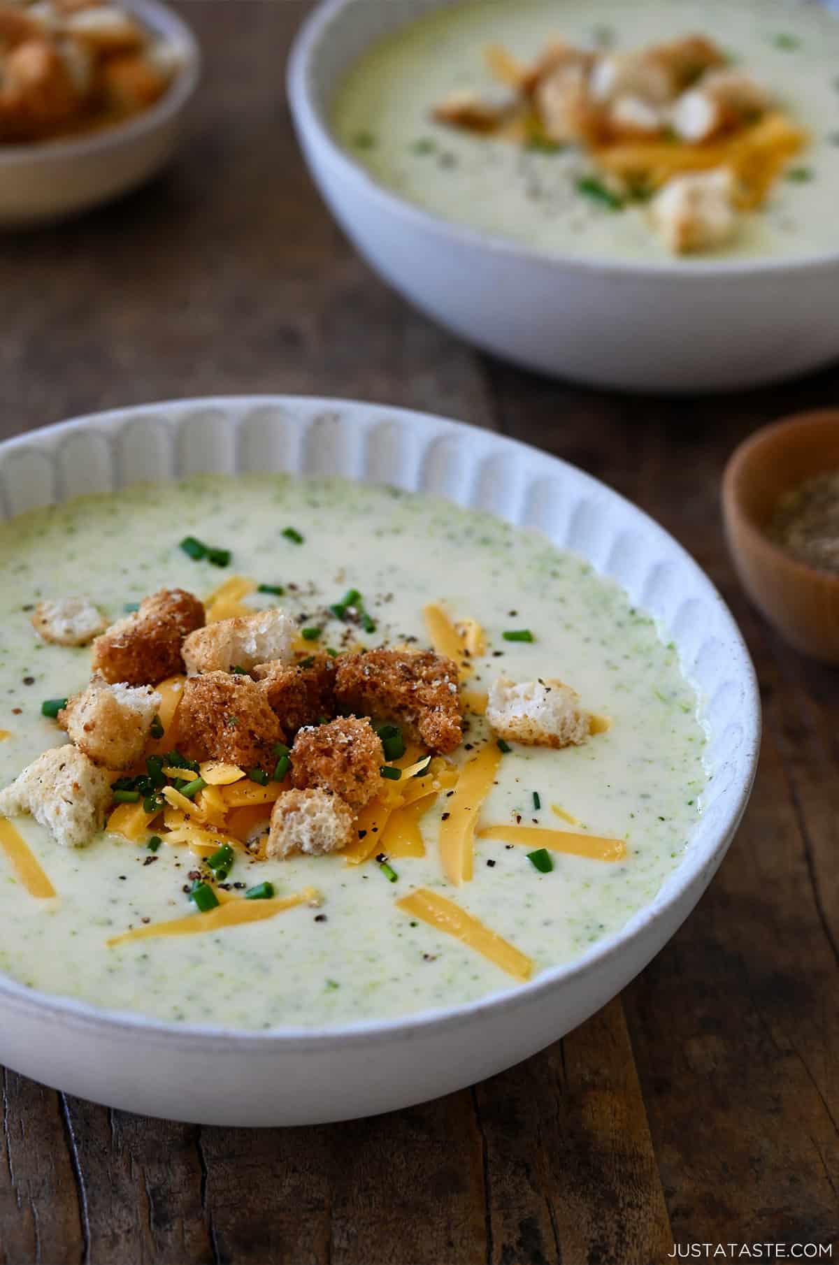 A bowl containing creamy homemade broccoli cheddar soup topped with homemade croutons, chopped chives and shredded cheddar cheese.