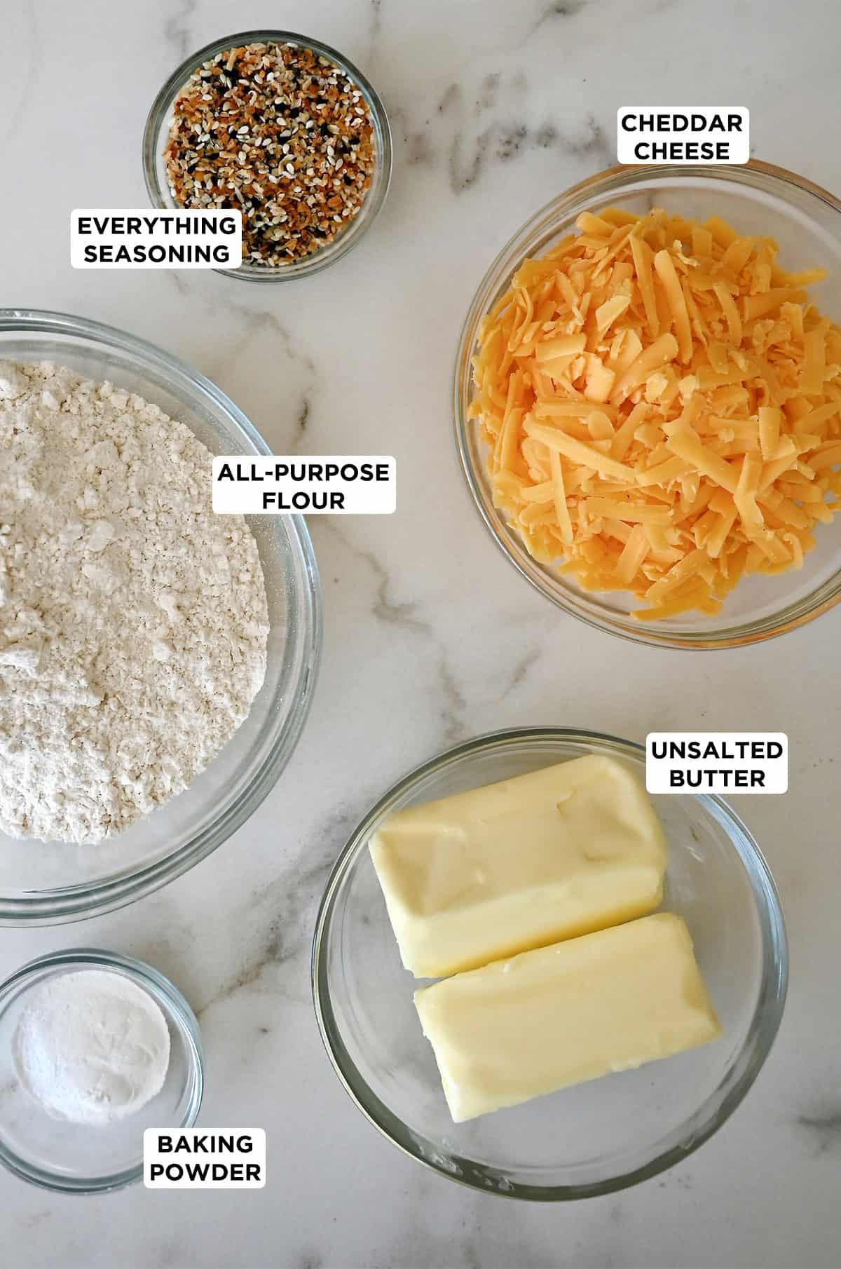 Cheese cookie ingredients in bowls, including cheddar cheese, flour, butter, baking powder and everything bagel seasoning.