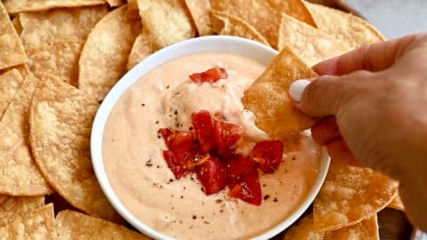 High-protein cottage cheese queso dip in a bowl topped with roasted tomatoes and black pepper on a plate surrounded by tortilla chips.