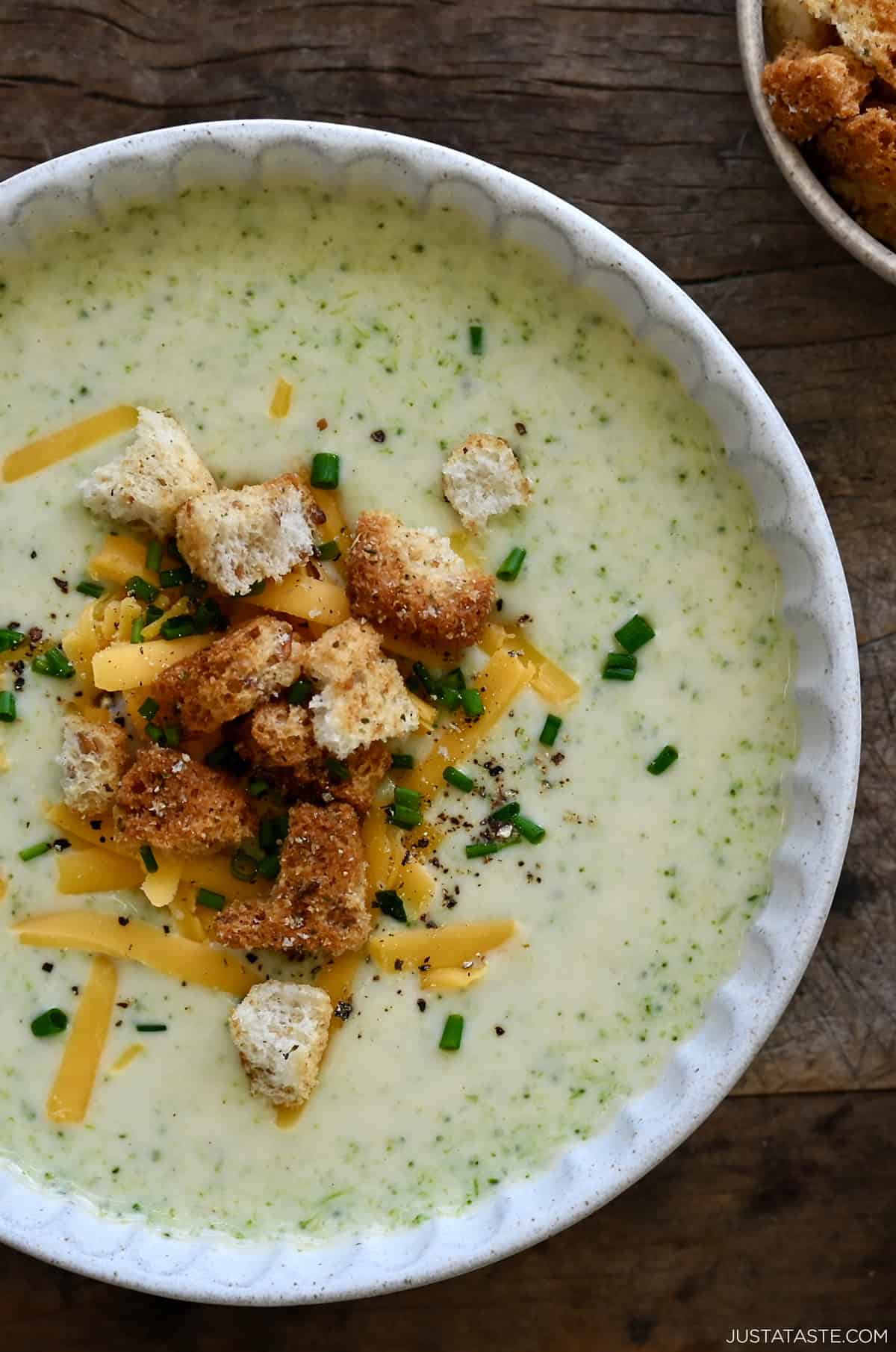 A bowl of creamy broccoli cheddar soup topped with croutons, shredded cheese and chopped chives.