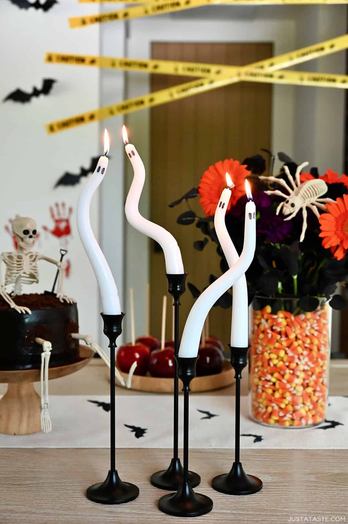 Four DIY ghost candles in black candlesticks on a Halloween-themed table with a skeleton cake.