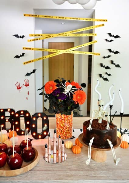 A Halloween-themed tablescape with a chocolate skeleton cake, an orange and black floral arrangement in a vase with candy corn, bat runner, melting finger candles, and mini pumpkins.
