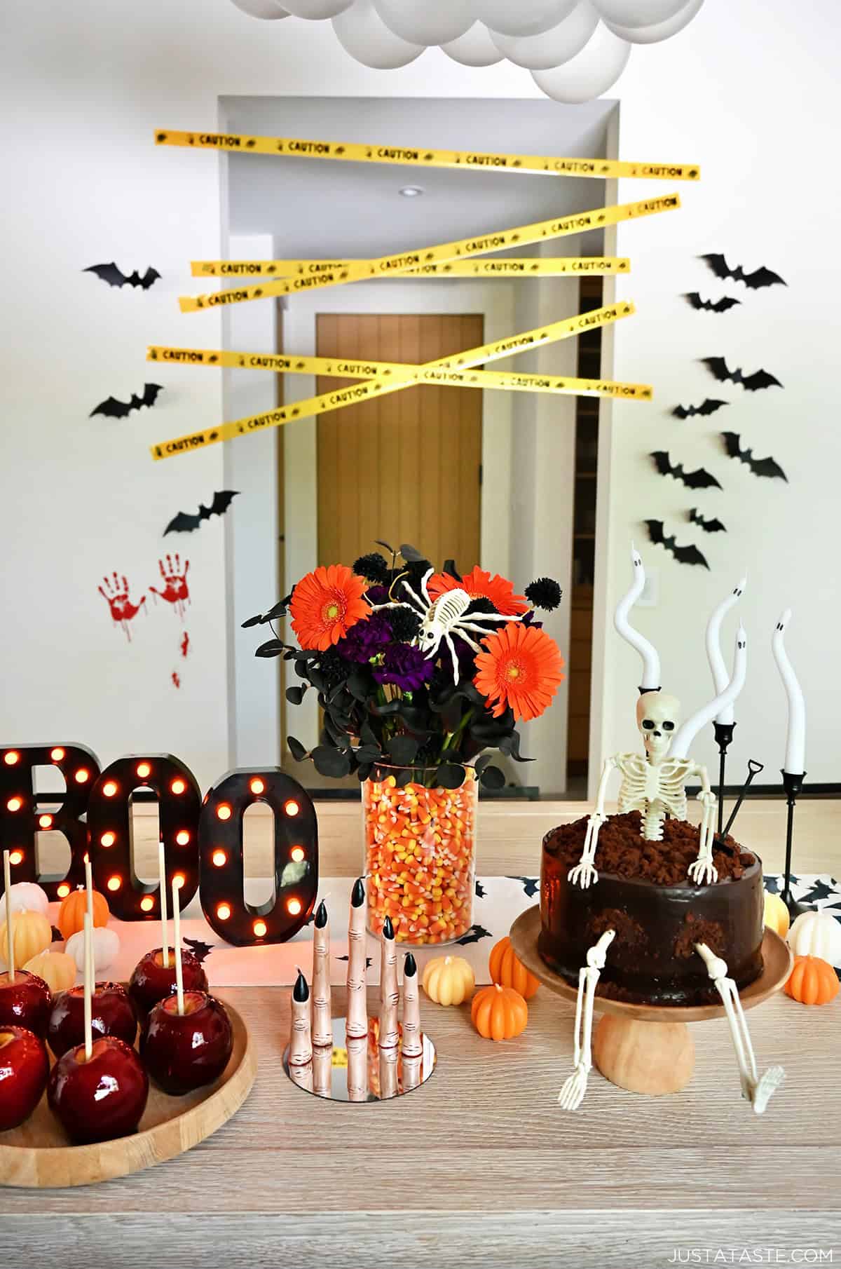 A Halloween-themed tablescape with a chocolate skeleton cake, an orange and black floral arrangement in a vase with candy corn, bat runner, melting finger candles, and mini pumpkins.