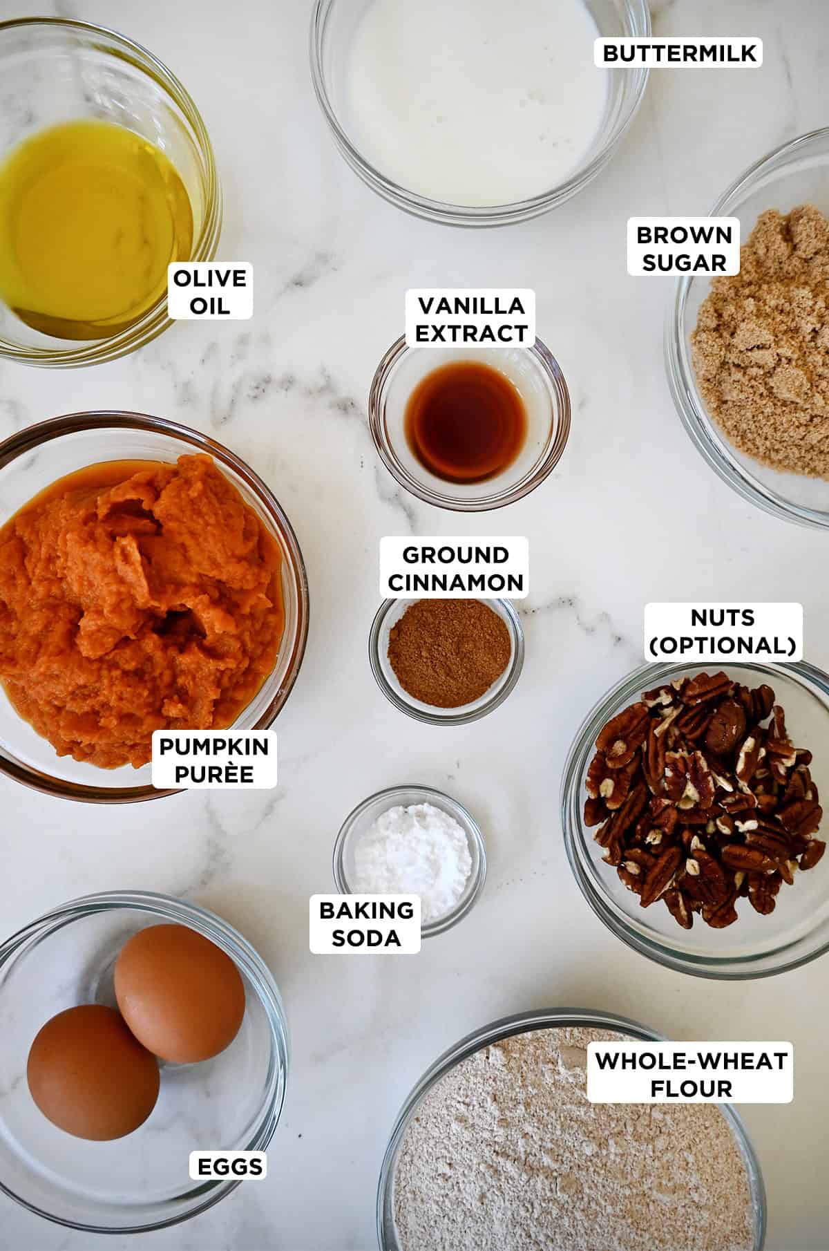 All the ingredients needed to make pumpkin muffins in various sizes of glass bowls, including pumpkin puree, olive oil, brown sugar, vanilla extract, whole wheat flour, buttermilk, eggs, baking soda, pumpkin pie spice and chopped pecans.