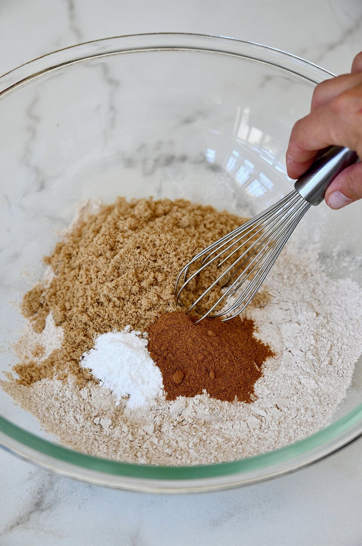 A glass bowl containing the dry ingredients for making healthy pumpkin muffins, including pumpkin pie spice, whole wheat flour, baking soda, salt and brown sugar.