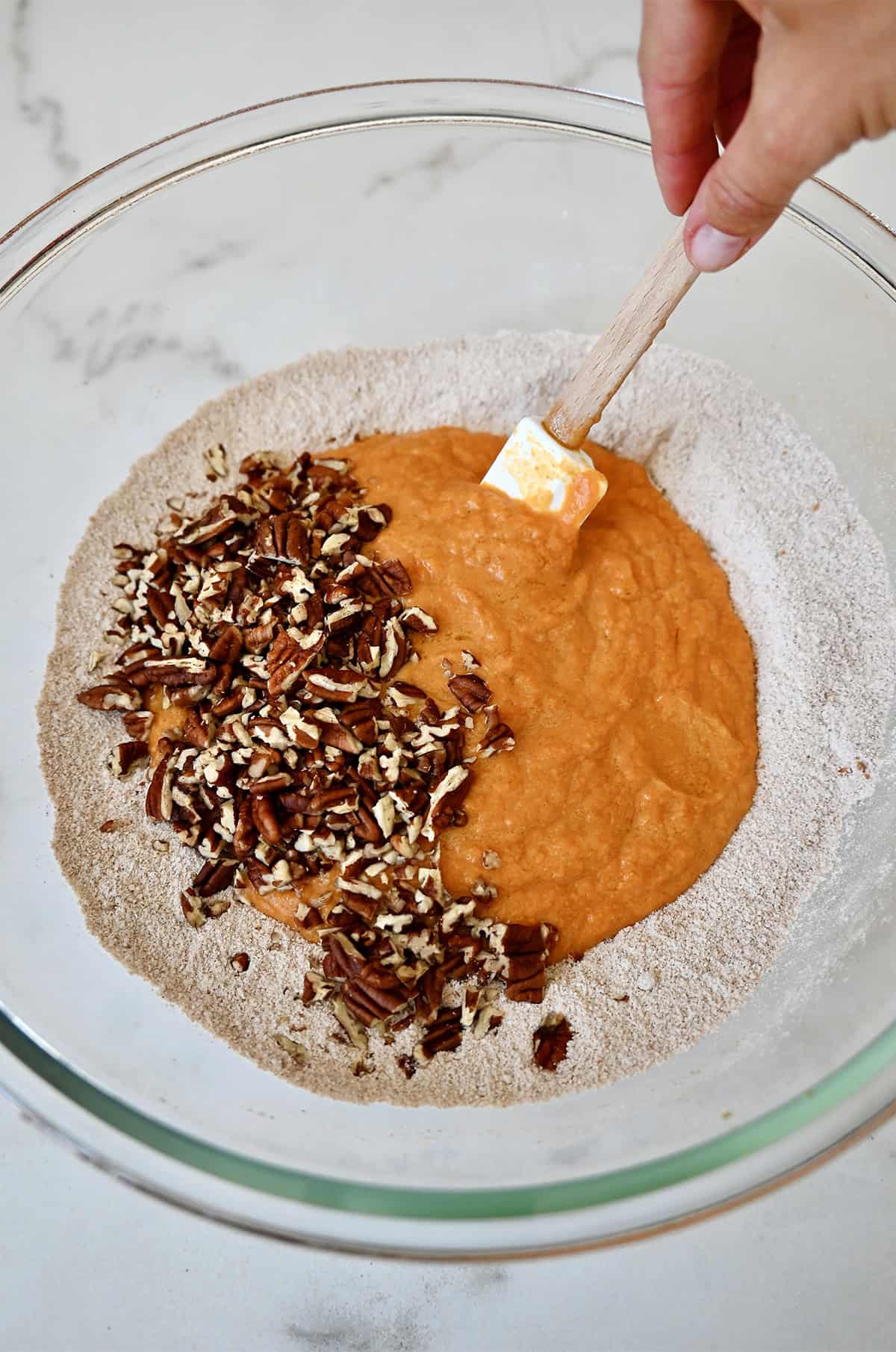 Folding chopped pecans into pumpkin muffin batter with wet and dry ingredients combined in a mixing bowl.