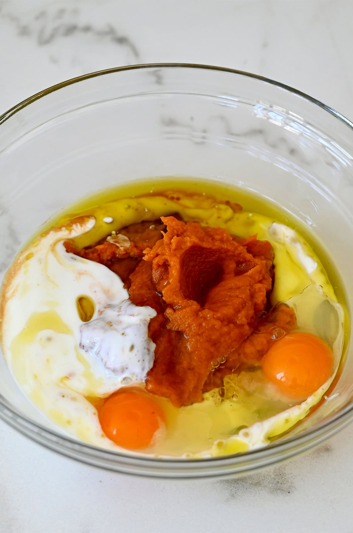 A glass bowl containing the wet ingredients for making pumpkin muffins, including pumpkin purée, eggs, buttermilk, vanilla extract and olive oil.