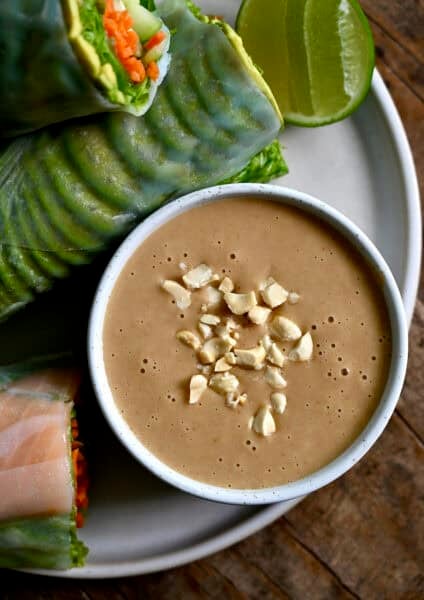 A bowl containing homemade Thai peanut sauce garnished with chopped peanuts on a plate with fresh spring rolls.