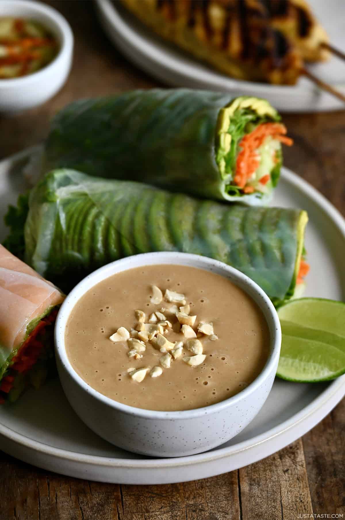 Thai peanut sauce in a bowl topped with chopped peanuts on a plate with fresh spring rolls.