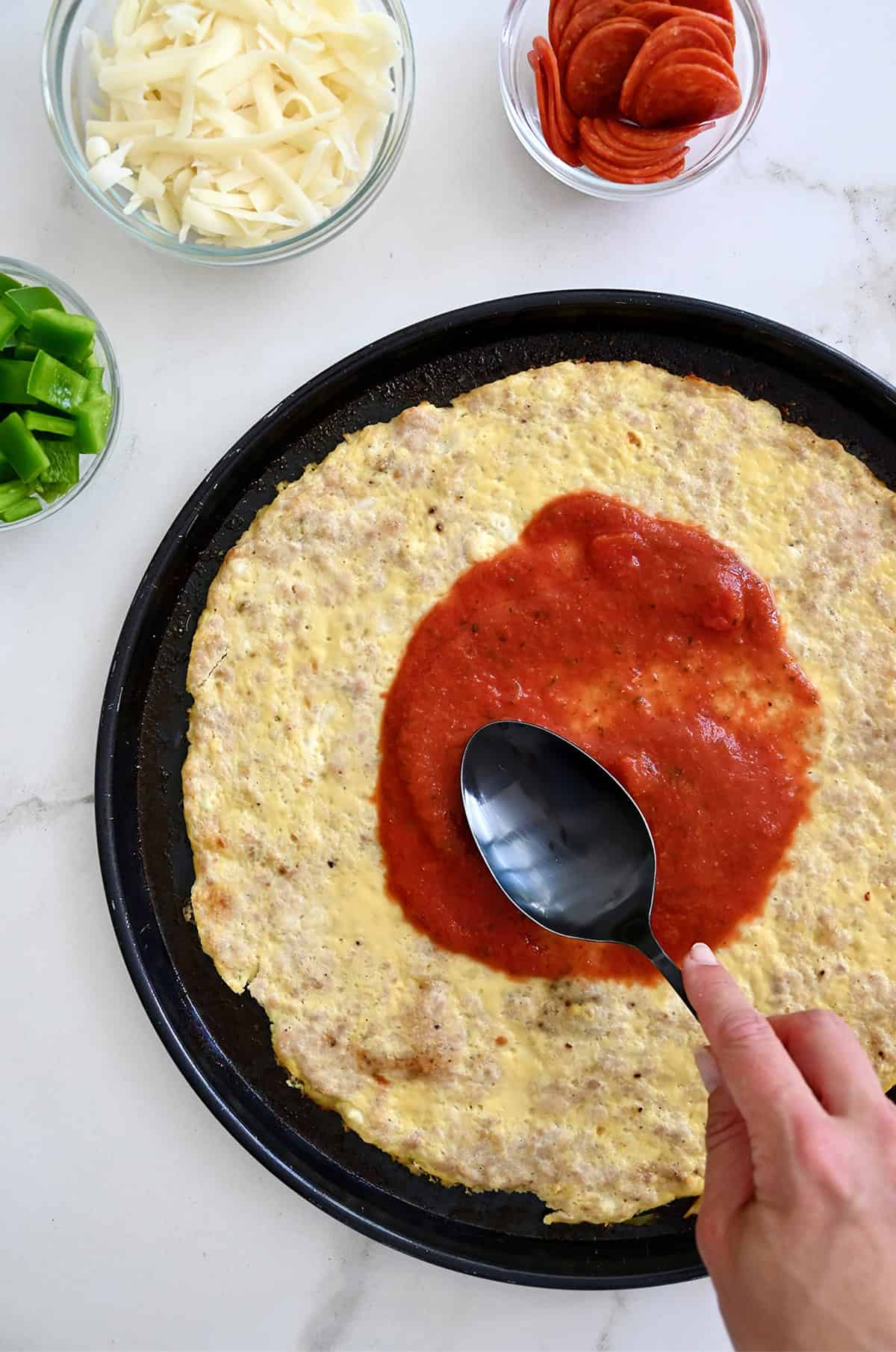 Using the back of a spoon to spread marinara sauce over a baked chicken crust pizza.