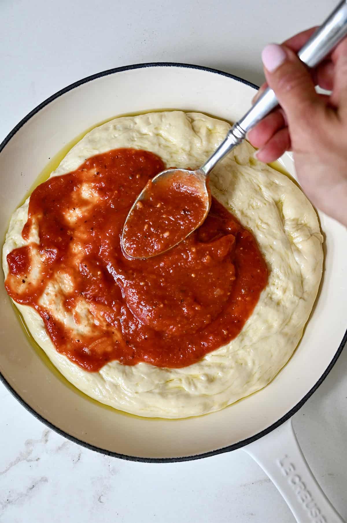 Spreading marinara sauce all the way to the edges of pizza dough in a cast iron pan.