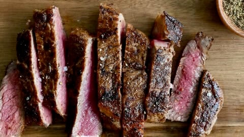 Sliced pan-seared steak on a wooden cutting board with small bowls containing salt and pepper nearby.