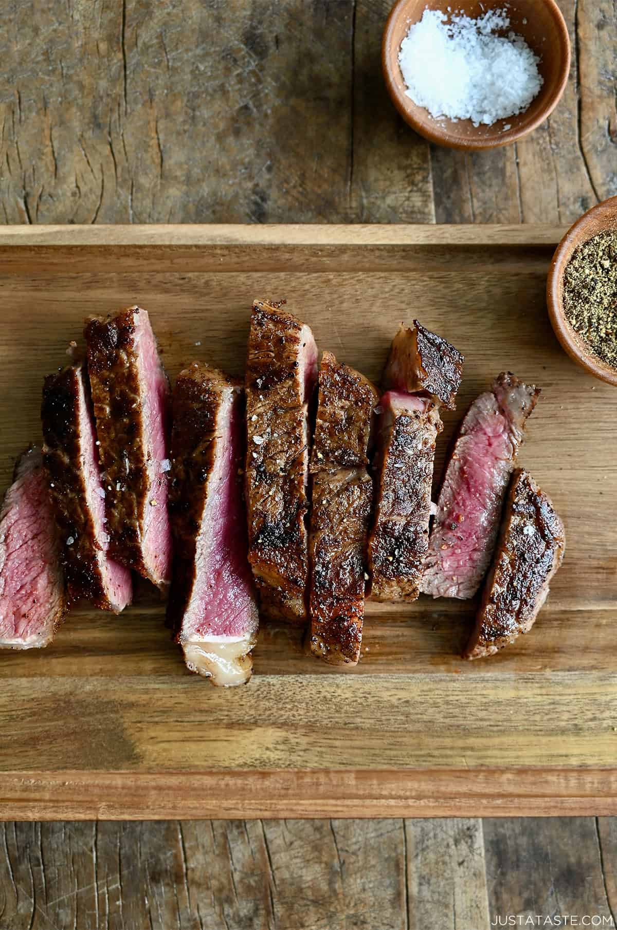 Sliced pan-seared steak on a wooden cutting board with small bowls containing salt and pepper nearby.