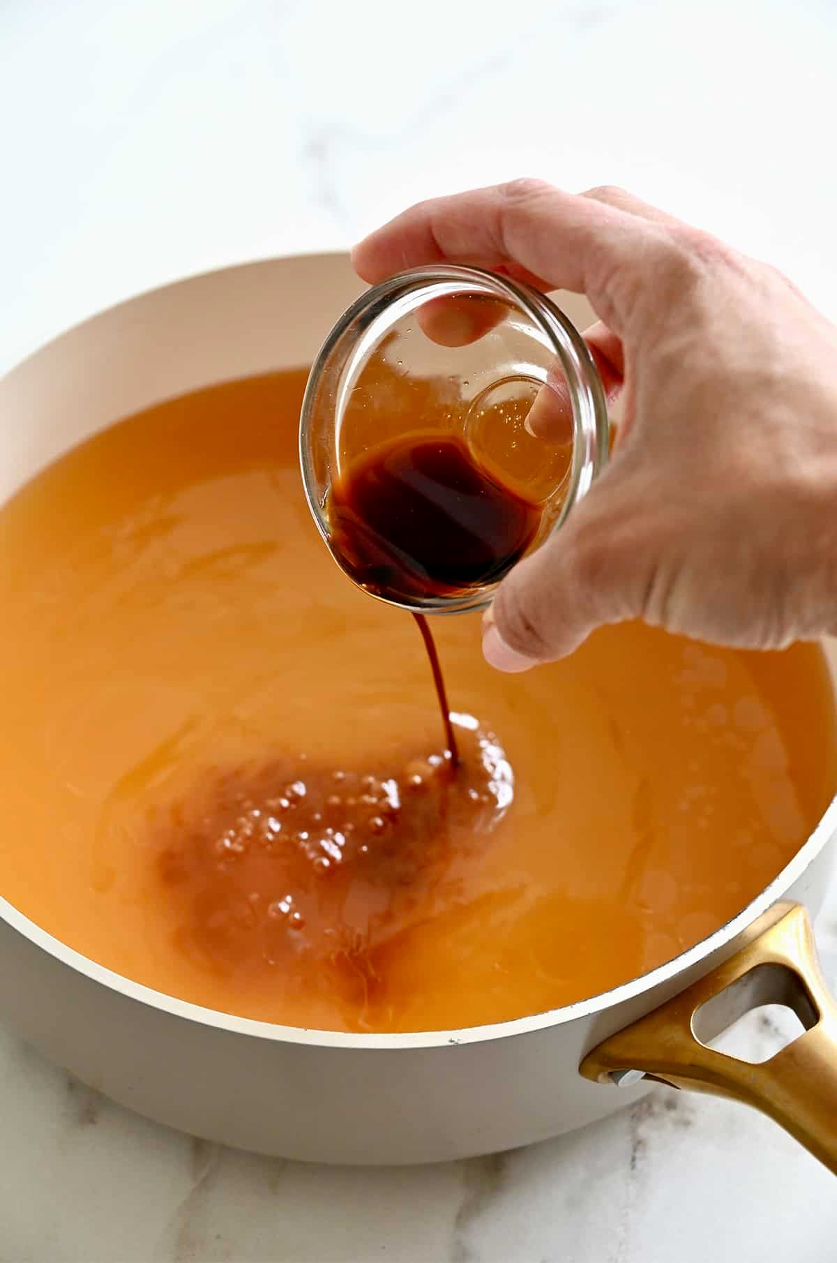 A mixture of soy sauce and sesame oil being poured from a small glass bowl into a stock pot containing chicken stock.