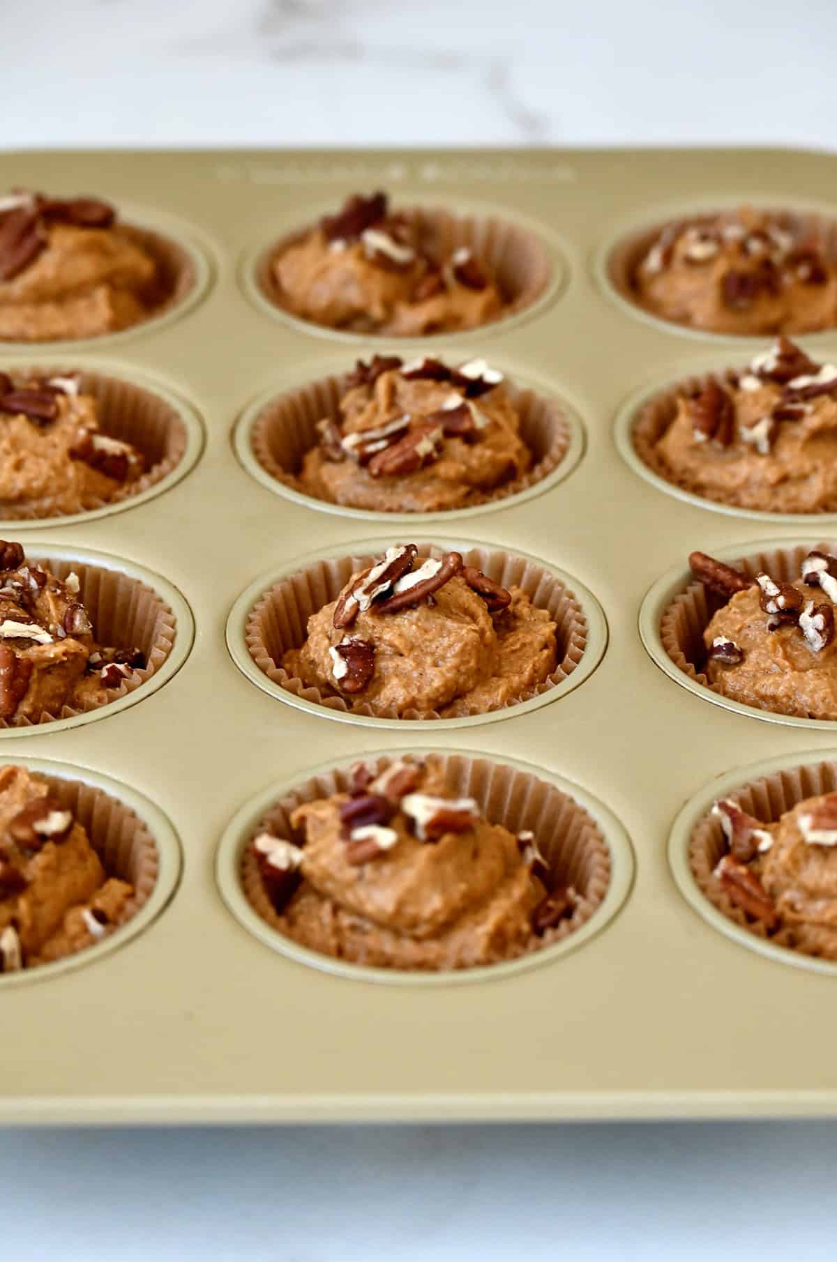 A muffin tin lined with baking cups, filled with healthy pumpkin muffin batter and topped with chopped pecans.