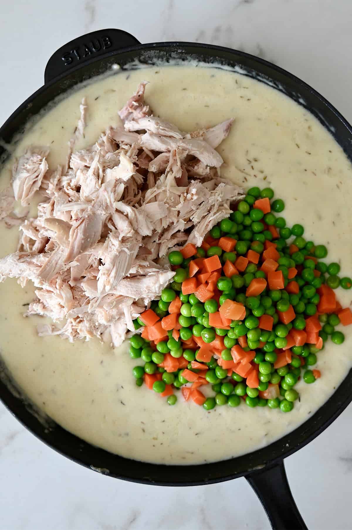 Shredded rotisserie chicken, peas and carrots being folded into creamy sauce in a skillet.
