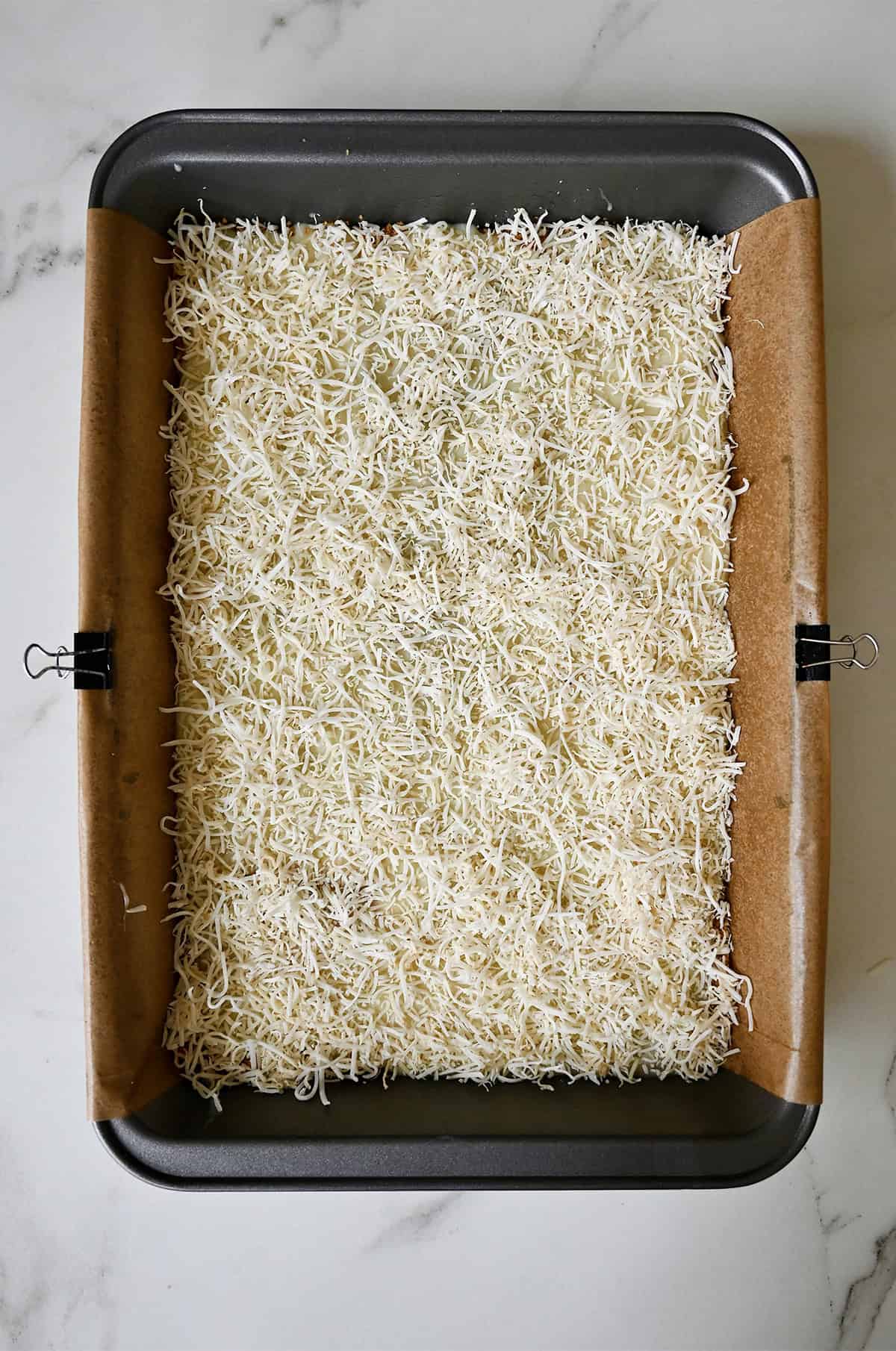 Shredded coconut atop a layer of sweetened condensed milk and a graham cracker base in a parchment paper-lined pan.